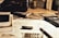 A workspace with technical drawings, a pen, a calculator, and a USB drive scattered on a desk. The setting suggests an environment focused on design or engineering, with blueprints and sketches partially visible. The lighting is warm and subdued.