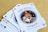 A fan of playing cards spread out on a light wooden table, featuring prominently is a Joker card with intricate illustrations. The Joker has a colorful jester hat and is holding a card and a hand of other cards.