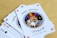 A fan of playing cards spread out on a light wooden table, featuring prominently is a Joker card with intricate illustrations. The Joker has a colorful jester hat and is holding a card and a hand of other cards.