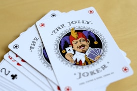 A fan of playing cards spread out on a light wooden table, featuring prominently is a Joker card with intricate illustrations. The Joker has a colorful jester hat and is holding a card and a hand of other cards.