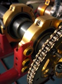 A close-up view of mechanical components featuring a chain and a series of cylindrical parts with golden and red structural elements. The chain is wound around a sprocket, and the parts are supported by a red frame with yellow bolts.