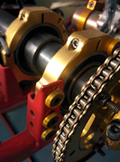 A close-up view of mechanical components featuring a chain and a series of cylindrical parts with golden and red structural elements. The chain is wound around a sprocket, and the parts are supported by a red frame with yellow bolts.