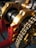 A close-up view of mechanical components featuring a chain and a series of cylindrical parts with golden and red structural elements. The chain is wound around a sprocket, and the parts are supported by a red frame with yellow bolts.