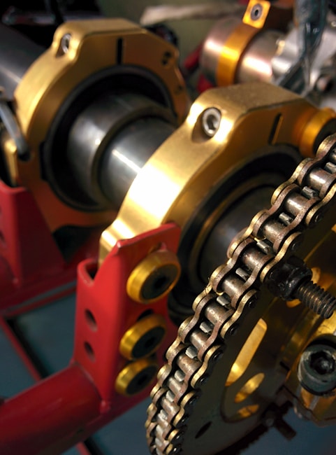 A close-up view of mechanical components featuring a chain and a series of cylindrical parts with golden and red structural elements. The chain is wound around a sprocket, and the parts are supported by a red frame with yellow bolts.