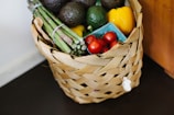A woven basket filled with fresh produce including avocados, asparagus, grape tomatoes, and a yellow bell pepper. The produce is arranged neatly, highlighting their vibrant colors against the natural tones of the basket.