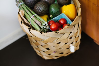 A woven basket filled with fresh produce including avocados, asparagus, grape tomatoes, and a yellow bell pepper. The produce is arranged neatly, highlighting their vibrant colors against the natural tones of the basket.