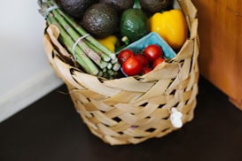 A woven basket filled with fresh produce including avocados, asparagus, grape tomatoes, and a yellow bell pepper. The produce is arranged neatly, highlighting their vibrant colors against the natural tones of the basket.