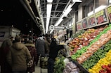 A busy outdoor market scene featuring a variety of colorful fruits and vegetables neatly arranged on display. Shoppers in warm clothing are browsing the produce under bright fluorescent lights at night. Signs with prices are visible above the produce. The background includes a store entrance and a postal truck.