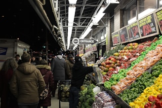 A busy outdoor market scene featuring a variety of colorful fruits and vegetables neatly arranged on display. Shoppers in warm clothing are browsing the produce under bright fluorescent lights at night. Signs with prices are visible above the produce. The background includes a store entrance and a postal truck.