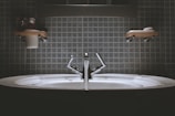 A bathroom sink with a modern design, featuring a shiny metallic faucet and a wide basin. Shelves above the sink hold toiletries such as a cup and soap dish. The wall is covered in square, blue tiles giving a clean and minimalist look.