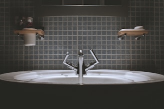 A bathroom sink with a modern design, featuring a shiny metallic faucet and a wide basin. Shelves above the sink hold toiletries such as a cup and soap dish. The wall is covered in square, blue tiles giving a clean and minimalist look.