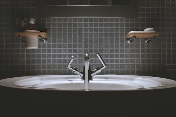 A bathroom sink with a modern design, featuring a shiny metallic faucet and a wide basin. Shelves above the sink hold toiletries such as a cup and soap dish. The wall is covered in square, blue tiles giving a clean and minimalist look.
