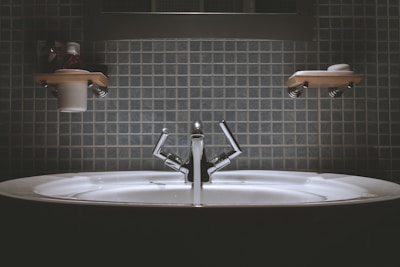 A bathroom sink with a modern design, featuring a shiny metallic faucet and a wide basin. Shelves above the sink hold toiletries such as a cup and soap dish. The wall is covered in square, blue tiles giving a clean and minimalist look.