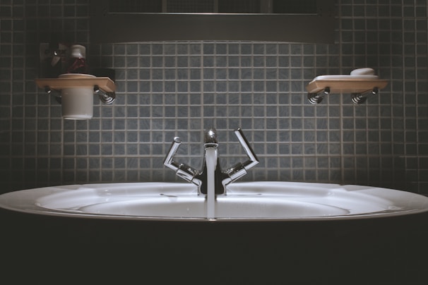A bathroom sink with a modern design, featuring a shiny metallic faucet and a wide basin. Shelves above the sink hold toiletries such as a cup and soap dish. The wall is covered in square, blue tiles giving a clean and minimalist look.