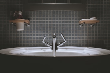 A bathroom sink with a modern design, featuring a shiny metallic faucet and a wide basin. Shelves above the sink hold toiletries such as a cup and soap dish. The wall is covered in square, blue tiles giving a clean and minimalist look.