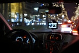 A nighttime scene from inside a car showing the dashboard and a GPS device mounted on the windshield. The dashboard is illuminated with glowing speedometers and electronics. Outside, a street is brightly decorated with blurred festive lights, and cars are visible ahead with their brake lights on.