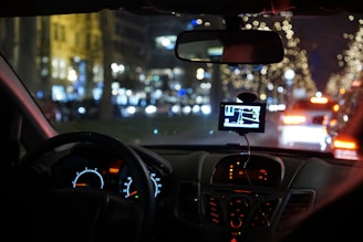 A nighttime scene from inside a car showing the dashboard and a GPS device mounted on the windshield. The dashboard is illuminated with glowing speedometers and electronics. Outside, a street is brightly decorated with blurred festive lights, and cars are visible ahead with their brake lights on.