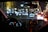 A nighttime scene from inside a car showing the dashboard and a GPS device mounted on the windshield. The dashboard is illuminated with glowing speedometers and electronics. Outside, a street is brightly decorated with blurred festive lights, and cars are visible ahead with their brake lights on.