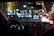 A nighttime scene from inside a car showing the dashboard and a GPS device mounted on the windshield. The dashboard is illuminated with glowing speedometers and electronics. Outside, a street is brightly decorated with blurred festive lights, and cars are visible ahead with their brake lights on.