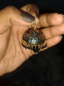 A hand is holding an intricately designed earring with a dome shape. The earring features a pattern of red and gold colors with black beads dangling around the edge. The fingernails have black glittery nail polish.