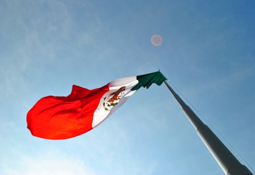 A large flag with distinct red, white, and green horizontal stripes, adorned with a detailed emblem in the center, billows in the wind against a clear blue sky. The flag is hoisted on a tall metal pole.