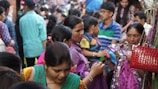A bustling market scene with a crowd of people engaged in shopping. Women and children are closely examining colorful fabrics and textiles. The atmosphere is lively and packed with people wearing vibrant clothing.