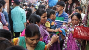 A bustling market scene with a crowd of people engaged in shopping. Women and children are closely examining colorful fabrics and textiles. The atmosphere is lively and packed with people wearing vibrant clothing.