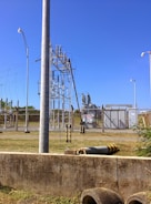 A fenced electrical substation with high voltage equipment and warning signs. There are several utility poles and transmission lines. The sky is clear and blue, and the surrounding area has grass and a few plants.