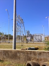 A fenced electrical substation with high voltage equipment and warning signs. There are several utility poles and transmission lines. The sky is clear and blue, and the surrounding area has grass and a few plants.