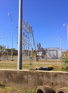 A fenced electrical substation with high voltage equipment and warning signs. There are several utility poles and transmission lines. The sky is clear and blue, and the surrounding area has grass and a few plants.