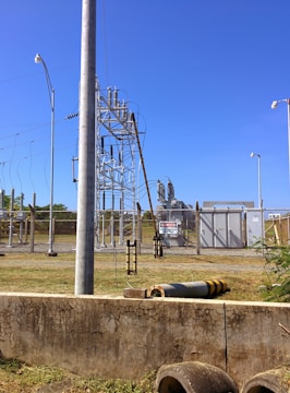 A fenced electrical substation with high voltage equipment and warning signs. There are several utility poles and transmission lines. The sky is clear and blue, and the surrounding area has grass and a few plants.