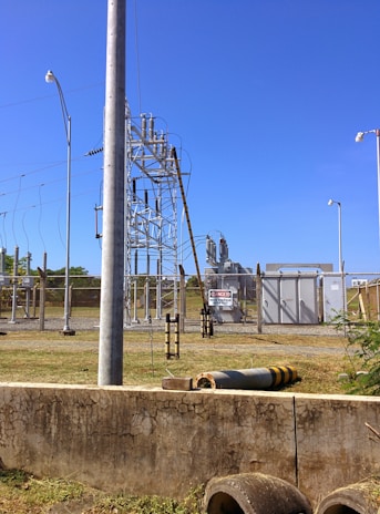 A fenced electrical substation with high voltage equipment and warning signs. There are several utility poles and transmission lines. The sky is clear and blue, and the surrounding area has grass and a few plants.