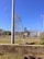 A fenced electrical substation with high voltage equipment and warning signs. There are several utility poles and transmission lines. The sky is clear and blue, and the surrounding area has grass and a few plants.