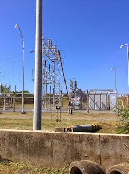 A fenced electrical substation with high voltage equipment and warning signs. There are several utility poles and transmission lines. The sky is clear and blue, and the surrounding area has grass and a few plants.