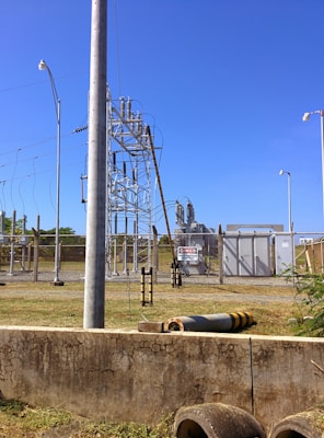A fenced electrical substation with high voltage equipment and warning signs. There are several utility poles and transmission lines. The sky is clear and blue, and the surrounding area has grass and a few plants.