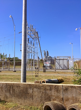 A fenced electrical substation with high voltage equipment and warning signs. There are several utility poles and transmission lines. The sky is clear and blue, and the surrounding area has grass and a few plants.