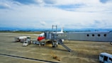 A commercial airplane with a red and white design is parked on an airport tarmac. It is connected to a passenger boarding bridge. Several service vehicles are nearby, including fuel and catering trucks. The scene is set against a backdrop of distant mountains beneath a cloudy sky.