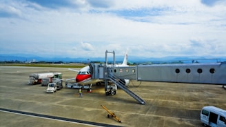 A commercial airplane with a red and white design is parked on an airport tarmac. It is connected to a passenger boarding bridge. Several service vehicles are nearby, including fuel and catering trucks. The scene is set against a backdrop of distant mountains beneath a cloudy sky.