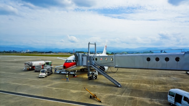 A commercial airplane with a red and white design is parked on an airport tarmac. It is connected to a passenger boarding bridge. Several service vehicles are nearby, including fuel and catering trucks. The scene is set against a backdrop of distant mountains beneath a cloudy sky.