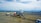 A commercial airplane with a red and white design is parked on an airport tarmac. It is connected to a passenger boarding bridge. Several service vehicles are nearby, including fuel and catering trucks. The scene is set against a backdrop of distant mountains beneath a cloudy sky.
