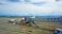 A commercial airplane with a red and white design is parked on an airport tarmac. It is connected to a passenger boarding bridge. Several service vehicles are nearby, including fuel and catering trucks. The scene is set against a backdrop of distant mountains beneath a cloudy sky.
