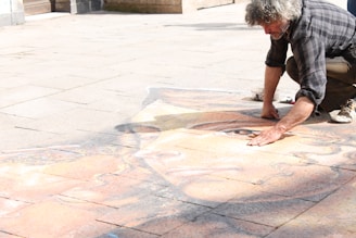 A man is kneeling on a paved surface while creating a detailed chalk drawing. The drawing appears to be a large, intricate portrait of a face, covering several pavement tiles. The artist is partially crouched, using one hand to add details to the artwork. Sunlight illuminates the scene, casting shadows on the ground.