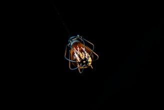 A single light bulb is encased in a protective metal cage, suspended against a dark, black background. The bulb emits a warm, yellow-orange glow, providing a stark contrast to the surrounding darkness.