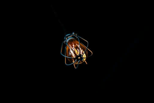 A single light bulb is encased in a protective metal cage, suspended against a dark, black background. The bulb emits a warm, yellow-orange glow, providing a stark contrast to the surrounding darkness.