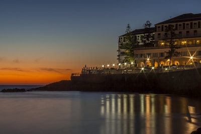A tranquil seaside view during sunset with a prominent building on the right featuring lit windows and lights creating starburst effects. The calm water in the foreground reflects the warm hues of the sky, transitioning from deep orange near the horizon to a dark blue overhead. Tall trees are silhouetted against the evening sky, adding to the serene atmosphere.