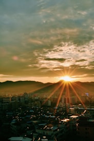 The image captures a cityscape at sunset, with the sun setting behind a range of hills. The sky is partially covered with clouds, and the sun's rays are visible, creating a serene and warm atmosphere. Buildings of various heights are seen in the foreground, illuminated by the soft golden light of the setting sun.