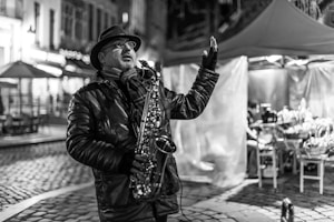 A street musician passionately playing a saxophone in a city setting at night. The man is wearing a hat, glasses, and a leather jacket. The background contains lit-up buildings and outdoor seating, along with cobblestone pavement.