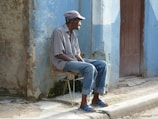 An elderly man is sitting on a makeshift chair beside a weathered blue wall. He wears a checkered shirt, blue jeans, sandals, and a flat cap. The setting appears to be a rustic street, with a wooden door behind him and a rough pavement underfoot.