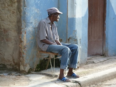 An elderly man is sitting on a makeshift chair beside a weathered blue wall. He wears a checkered shirt, blue jeans, sandals, and a flat cap. The setting appears to be a rustic street, with a wooden door behind him and a rough pavement underfoot.