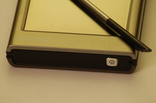A GPS device accompanied by a stylus is positioned on a flat surface. The device has a metallic casing with a power button visible at the bottom right corner. The stylus, which appears to be black, is resting diagonally across the device.